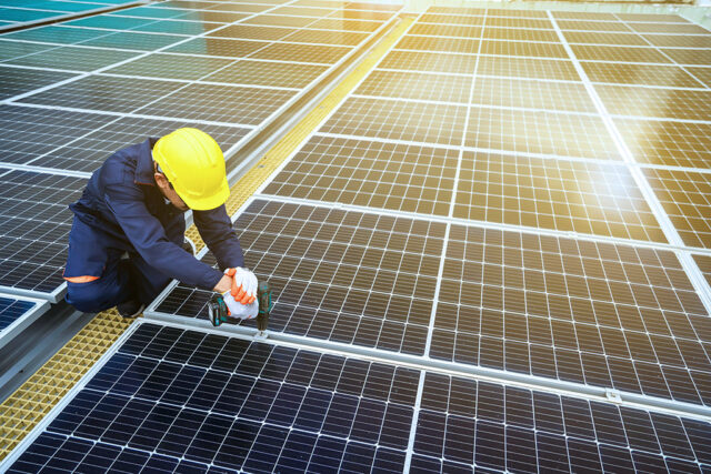 Eine Dachfläche voll spiegelnder Photovoltaik-Panele, ein Mann in Arbeitskleidung befestigt ein Panel mit dem Akkuschrauber